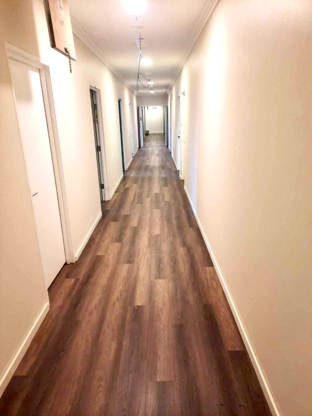 Long hostel corridor lined with doors, fitted with dark brown vinyl plank flooring — Drifters Hostel Byron Bay